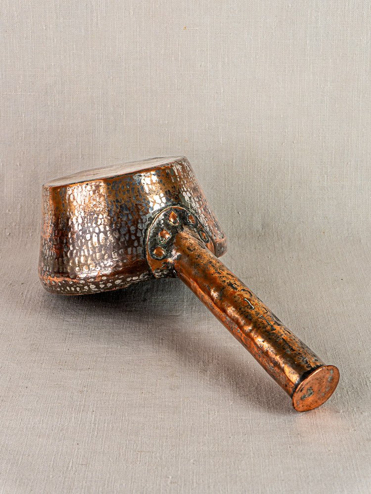 Antique French hand-forged copper saucepan with long handle and hammered finish on neutral fabric.