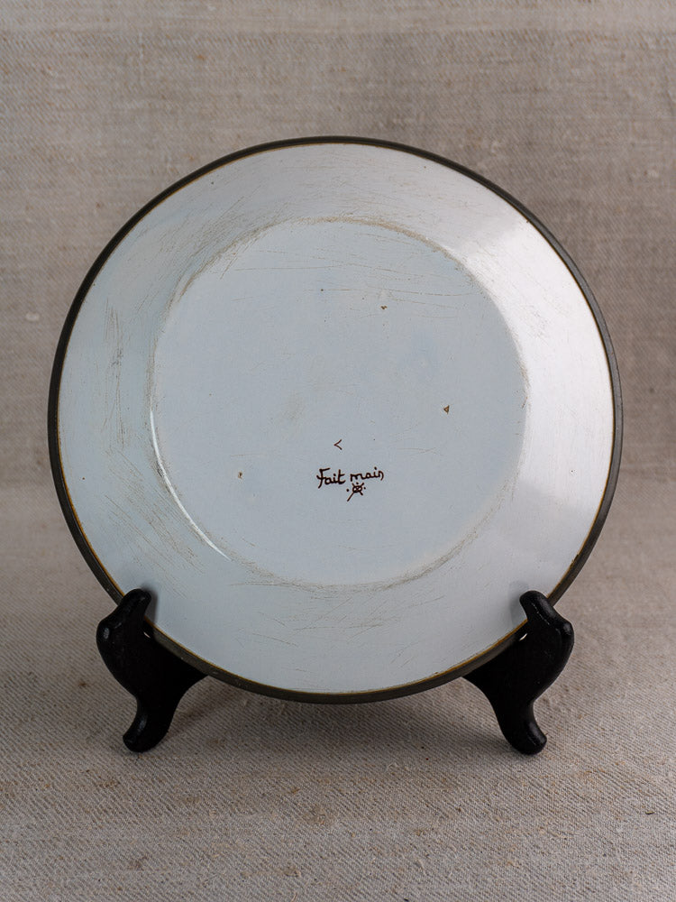 French faience wall plate, Moustiers style, black rim, “Fait main,” on beige textured surface.