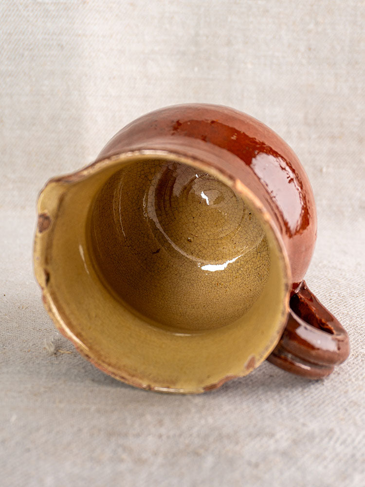 19th-century French earthenware pichet with brown glaze, yellow interior, and a small rim chip on beige textured fabric.