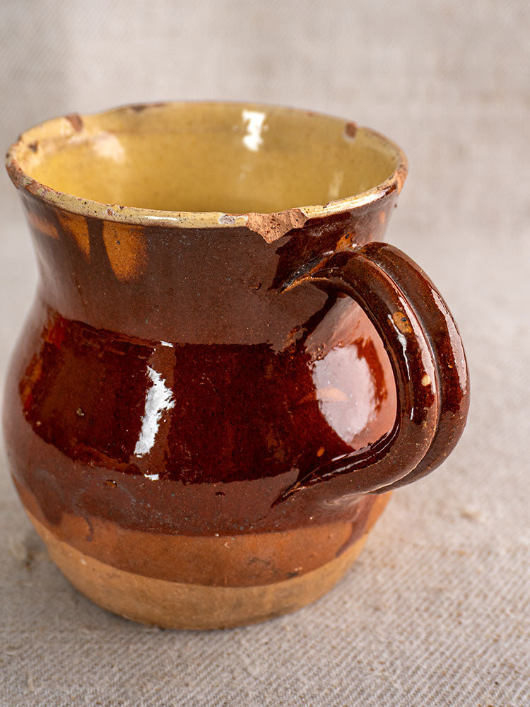 19th-century French pichet in rustic glazed earthenware with curved handle, yellow interior, and rim chips on textured fabric.