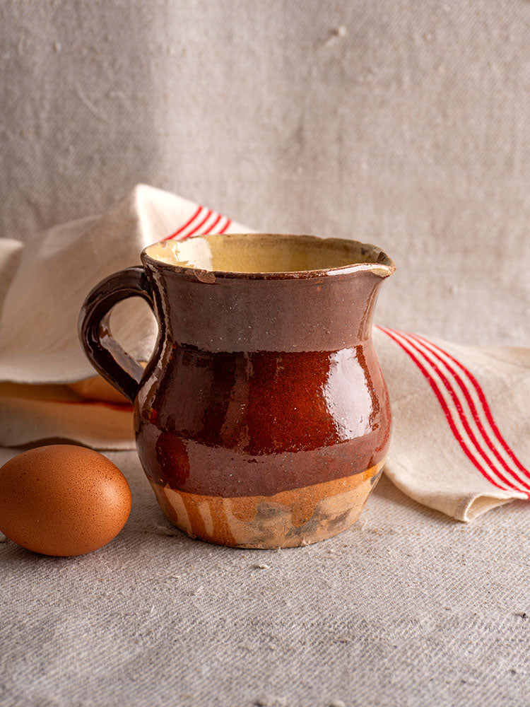 19th century French glazed earthenware pichet in rustic brown beside an egg on gray fabric, red-striped cloth folded behind.