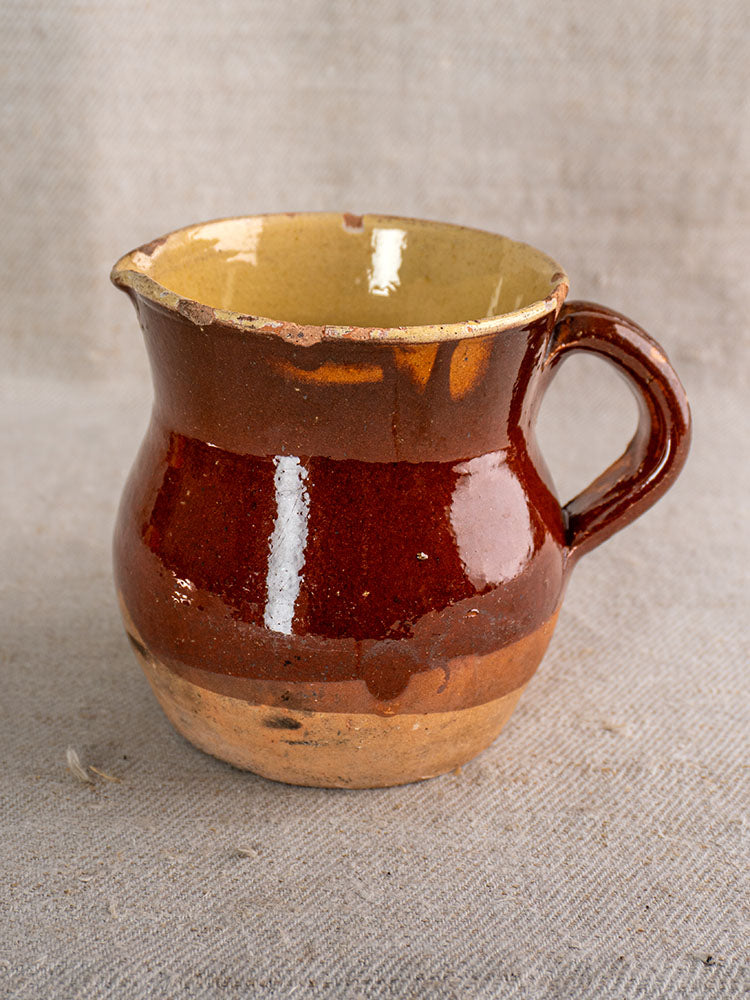 19th-century French Pichet, rustic half-dipped glazed earthenware pitcher on textured beige fabric background.