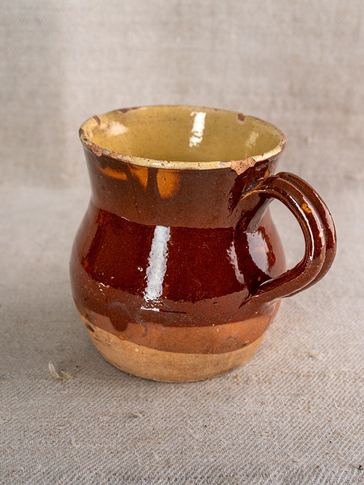 Rustic 19th-century French earthenware pichet with half-dipped glaze, wide base, and chipped rim on beige textured fabric.