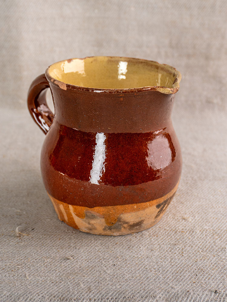 19th-century French earthenware pichet with brown glaze, cream interior, and single handle on textured beige fabric.