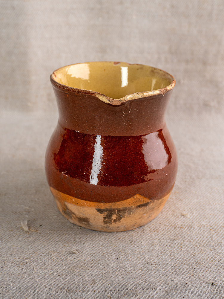 19th century French earthenware pichet with glossy brown glaze, rustic unglazed base, on coarse beige fabric.