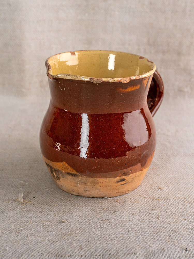 19th-century French glazed earthenware pichet with rustic half-dipped design and chipped rim on beige textured fabric.