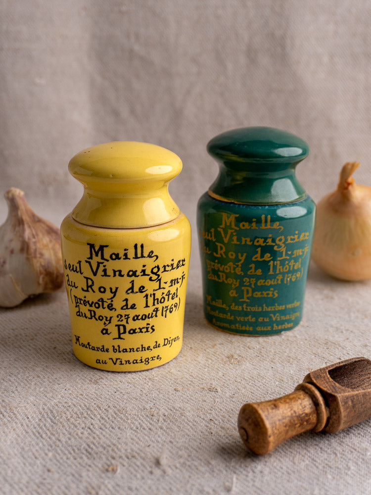 Pair of vintage French Maille mustard pots in teal and yellow with script, styled on textured cloth with garlic and cork.
