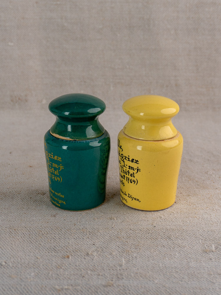 Pair of vintage French Maille mustard pots in teal and yellow with gold lettering, on beige fabric.