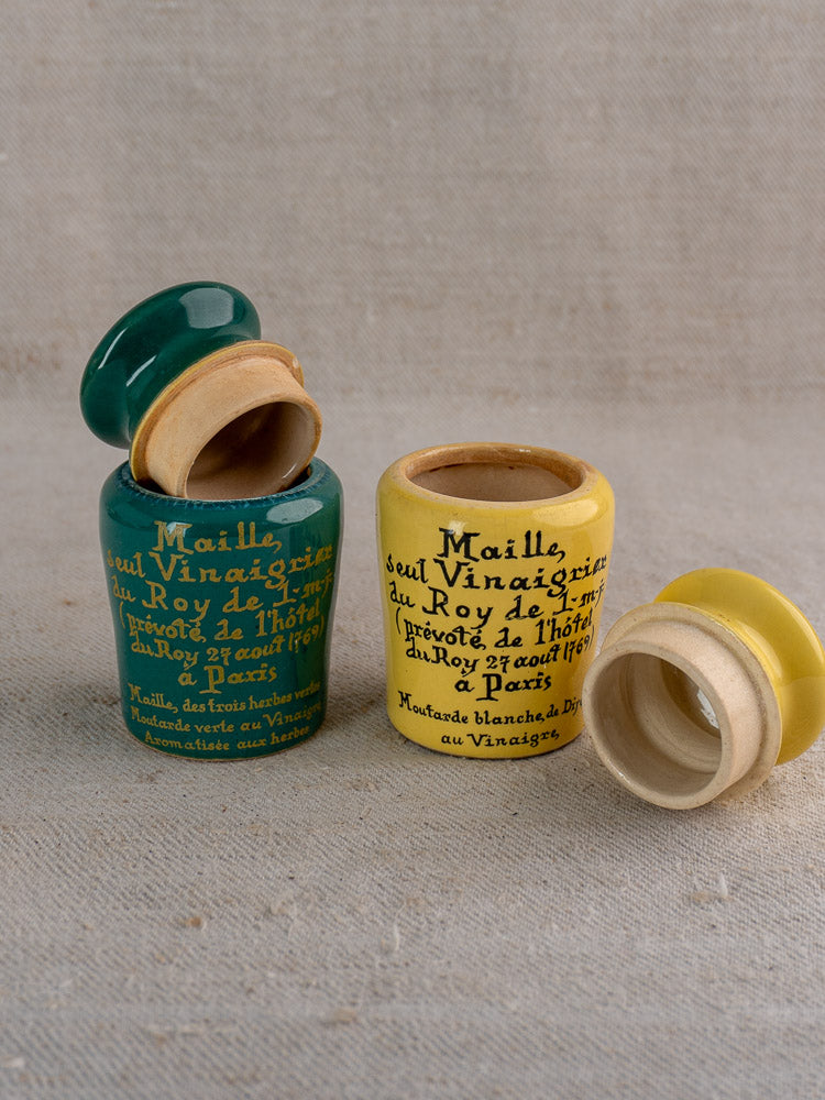 Pair of vintage French Maille mustard pots, teal and yellow, with handwritten labels and separate lids on beige fabric.