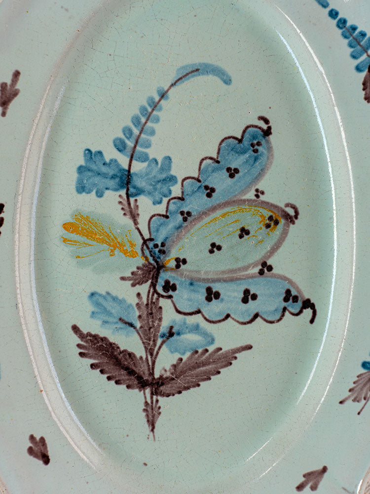Antique French faience Cul Noir platter with hand-painted blue and yellow butterfly amid blue and brown foliage on pale ground.