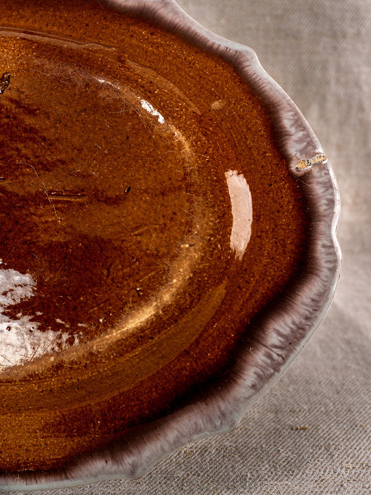 Antique French Cul Noir faience platter, late 18th century, glossy brown glaze, wavy rim, minor chip; on beige textured fabric.