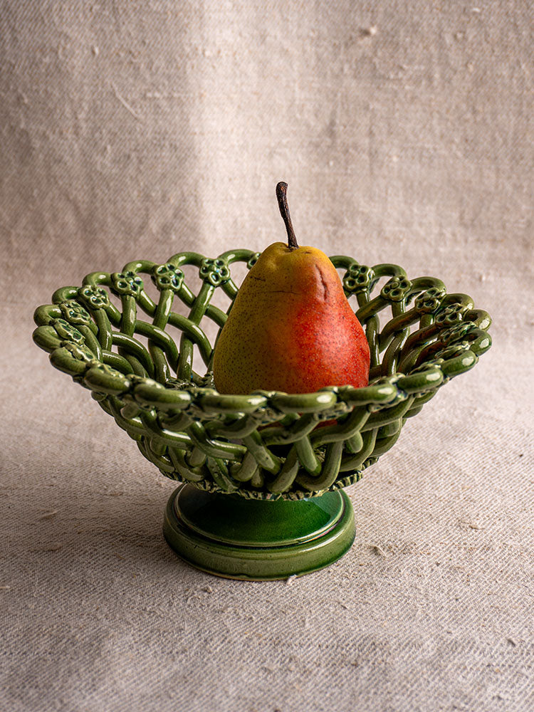 Vintage French Pichon Uzès green glazed woven basket holding a ripe pear, on textured beige fabric.