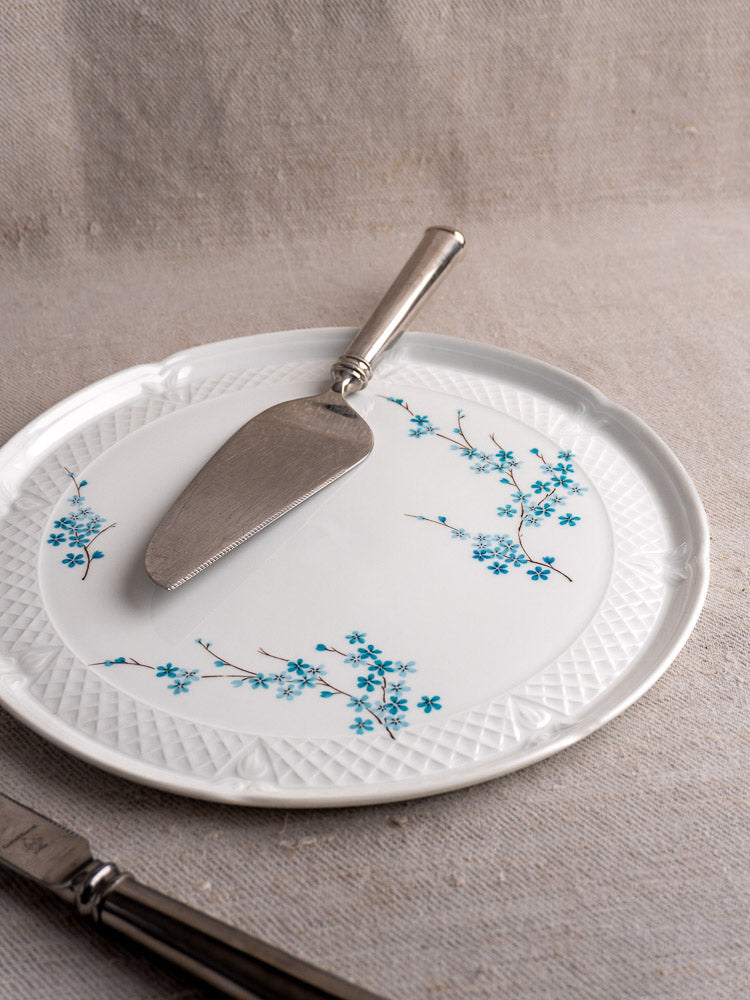 Vintage French Limoges porcelain cake plate with blue floral design, on beige cloth, metal cake server and knife handle nearby.