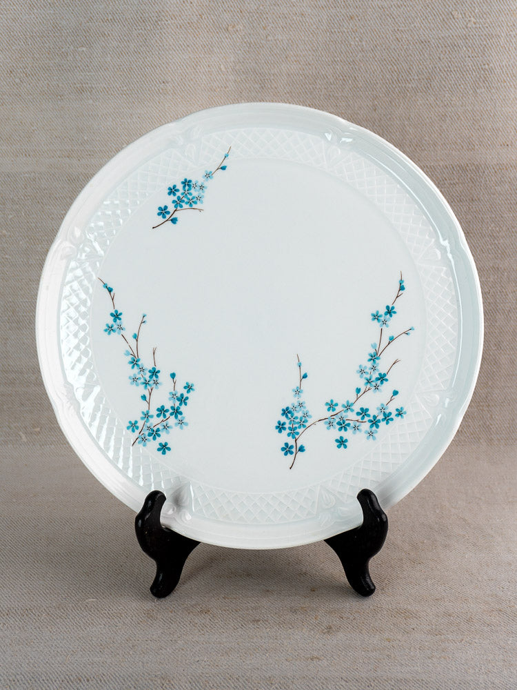 Vintage French Limoges porcelain cake plate with textured rim and blue floral motif on a black stand against beige fabric.