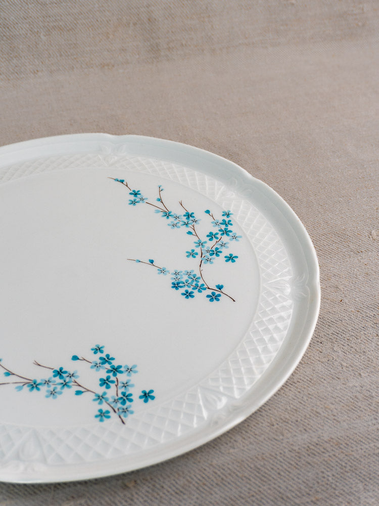 Vintage French Limoges porcelain cake plate with textured rim, blue floral pattern, on beige fabric surface.