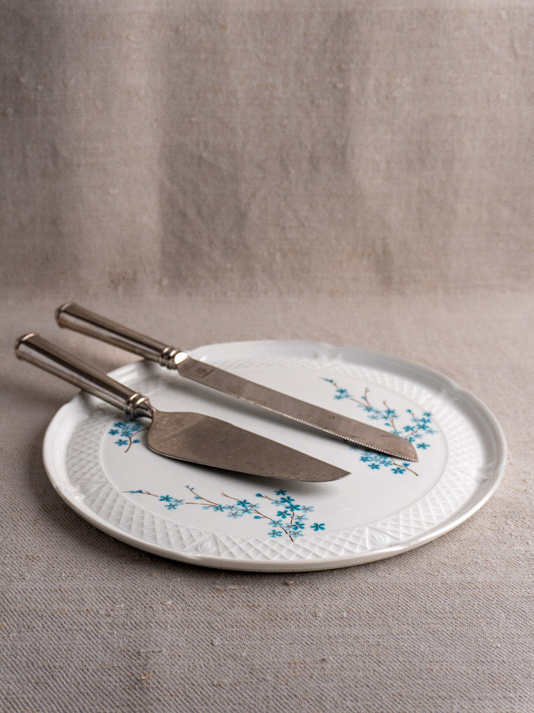 Vintage French Berry Limoges porcelain cake plate with blue floral motif, set on white plate over beige textured fabric.