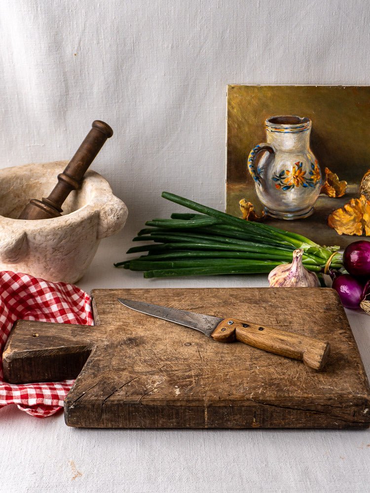 Antique 19th-century French hardwood cutting board with knife, vegetables, mortar and pestle, and red checkered cloth.