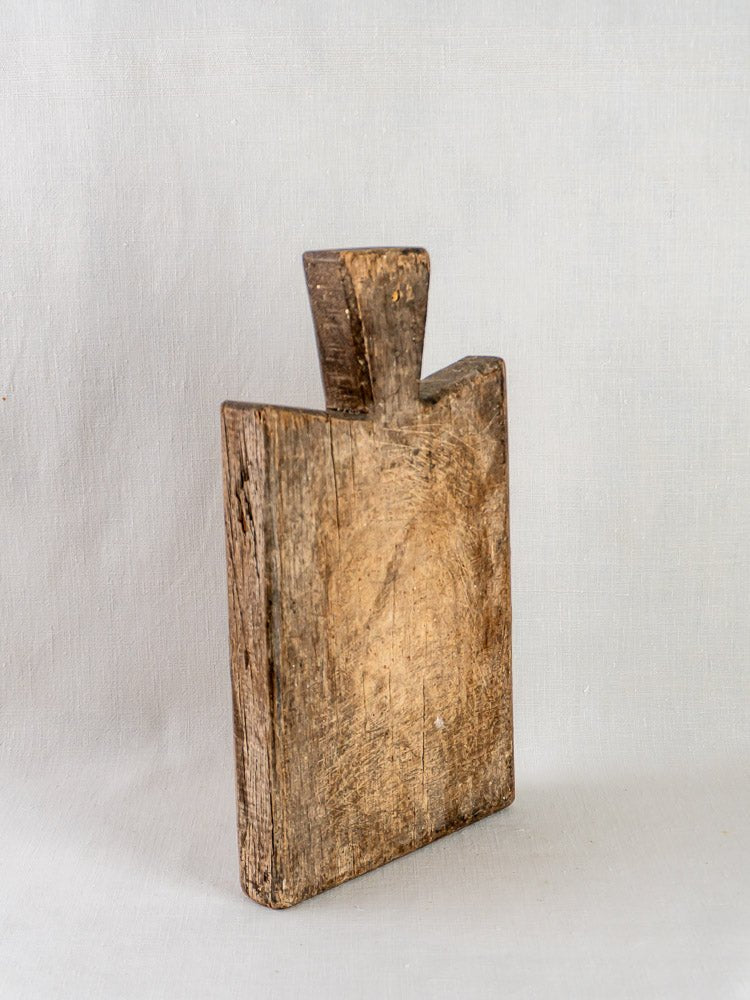 Antique 19th-century French hardwood cutting board with handle, upright on light gray surface, aged wood grain visible.