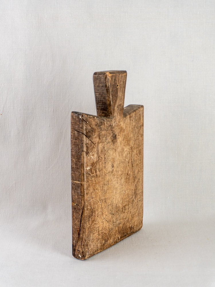 Antique 19th-century French hardwood cutting board with handle, rustic patina, upright on plain backdrop.