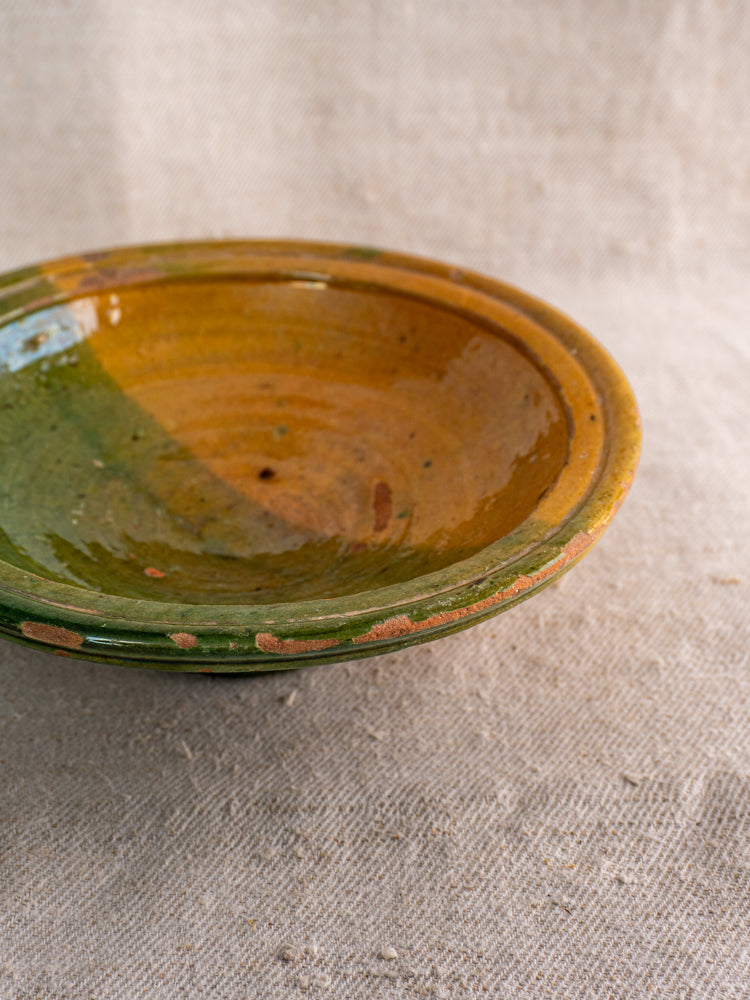 Antique French Provençal compotier with glossy green and ochre glaze, exposed clay rim, on textured beige fabric.