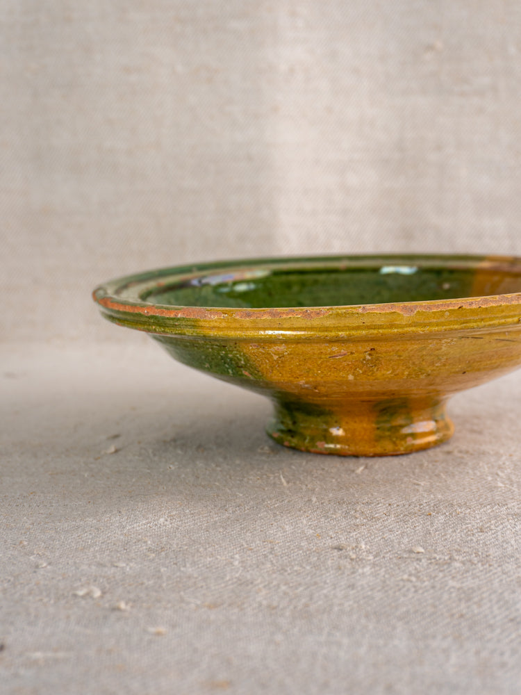 Antique French Provençal compotier with green and ochre glaze, uneven rim, on beige textured fabric.