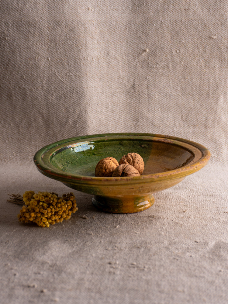 Antique French Provençal compotier with green and ochre glaze, filled with walnuts, on textured beige fabric.