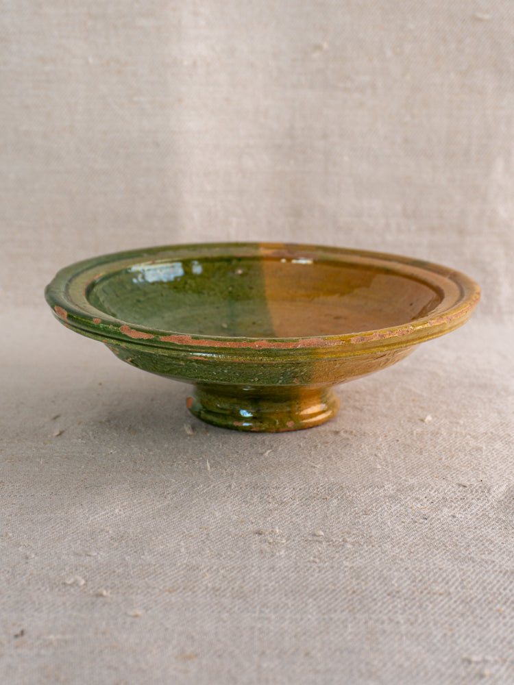 Antique French Provençal compotier with green and ochre glaze, brown flecks, on textured beige fabric background.