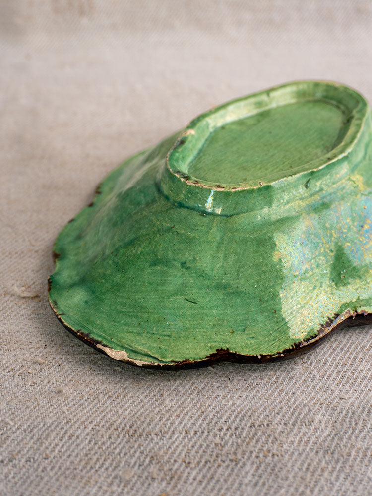 Antique French green glazed lobed coupelle, Dieulefit style, scalloped edges chipped to reveal brown beneath, on beige fabric.