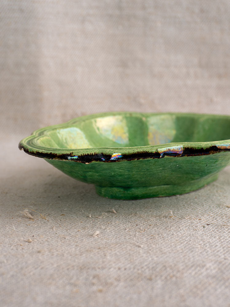 Antique French Dieulefit green glazed oval lobed dish with worn dark rim on textured beige fabric surface.