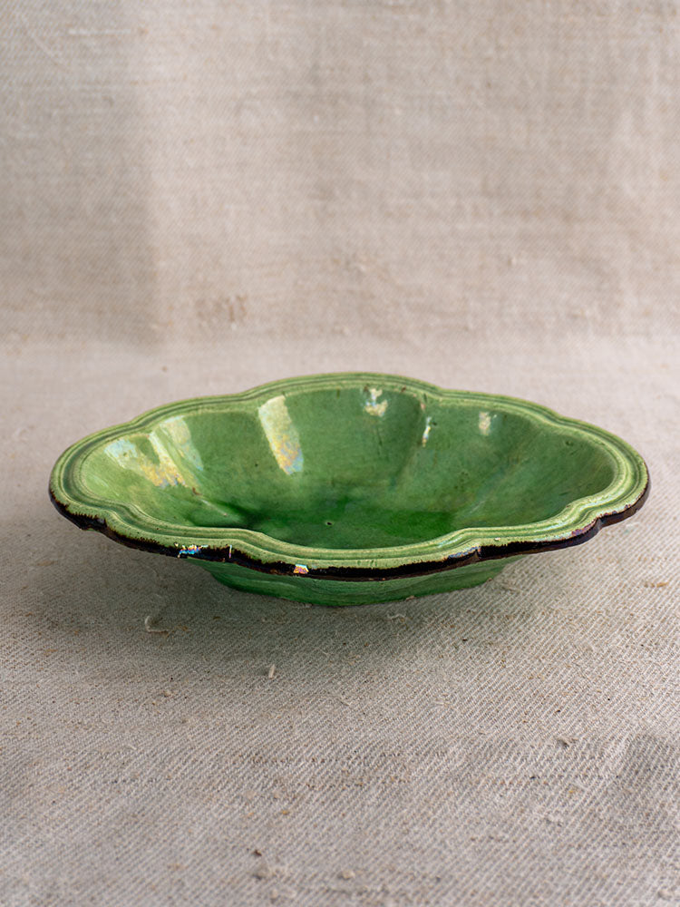 Antique French green glazed lobed oval coupelle with dark rim, likely Dieulefit, on textured beige fabric.