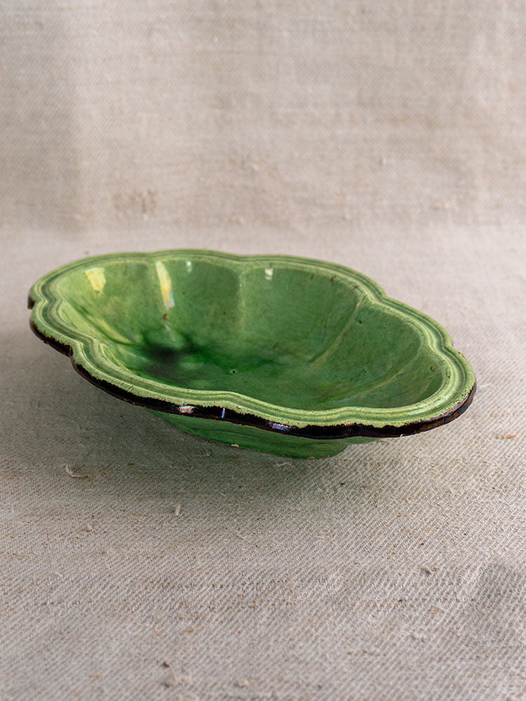 Antique French green glazed lobed oval dish with glossy scalloped edge on textured beige fabric.