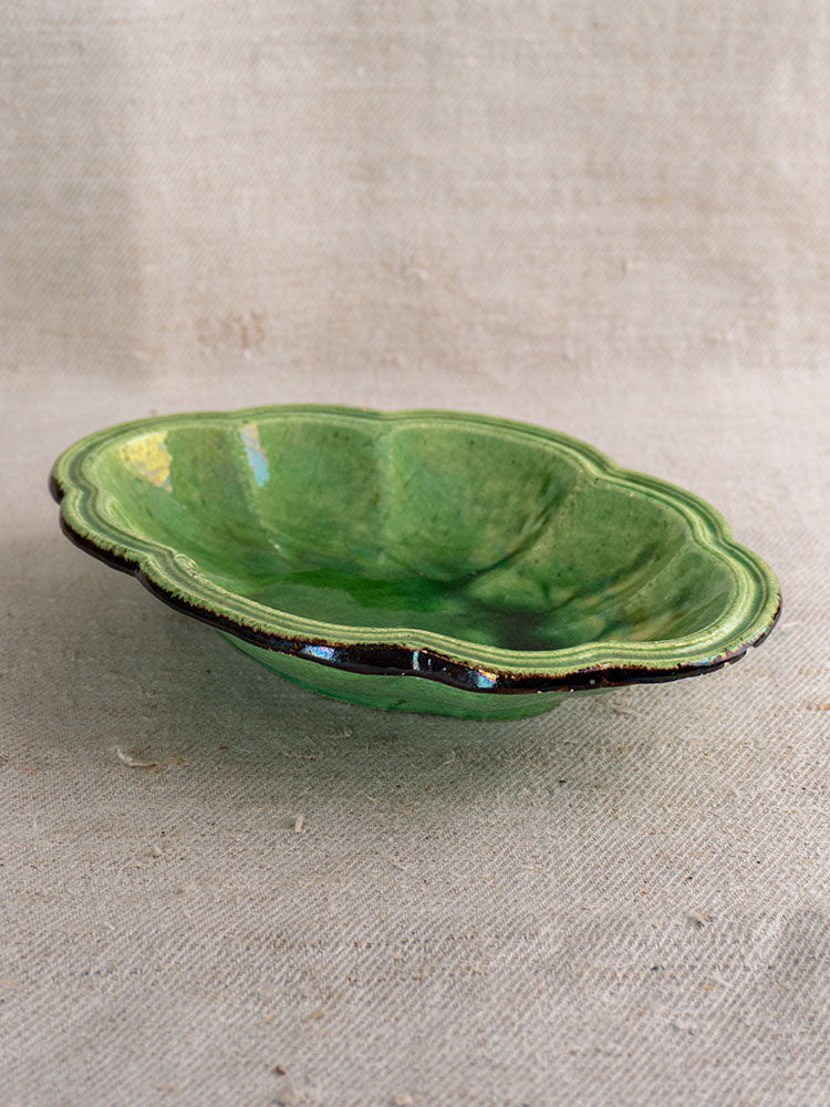 Antique French green glazed coupelle with scalloped edges and dark rim on textured beige fabric.