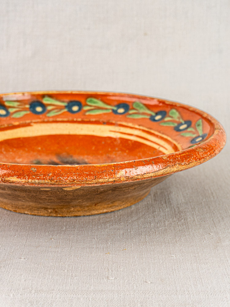 Antique French Alsatian Soufflenheim slipware plate with orange glaze, blue floral and green leaf border, on neutral fabric.