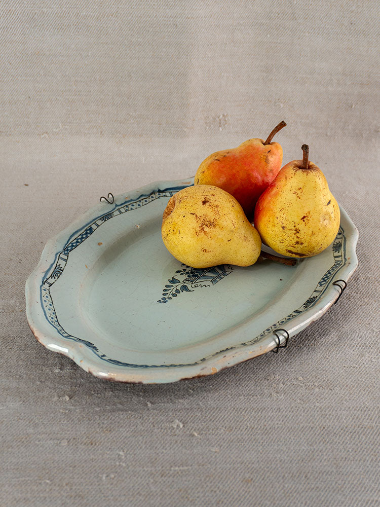 19th-century French faience platter, Rouen Cul Noir, blue edging, visible cracks; pears on neutral fabric background.