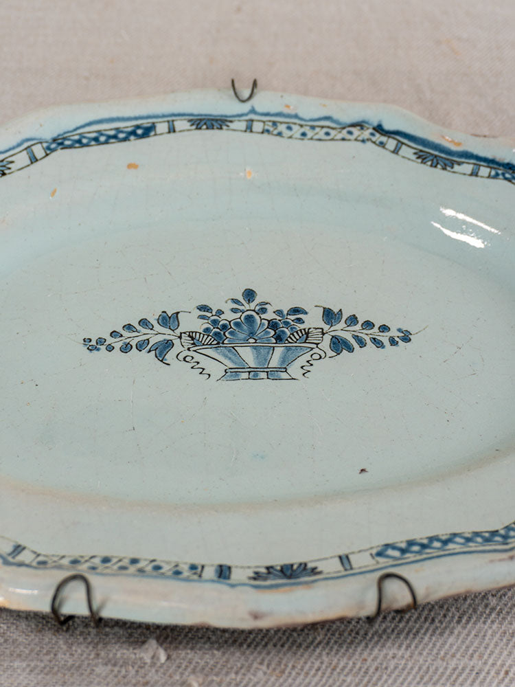 19th-century French faience platter, Rouen Cul Noir, blue floral basket motif, on beige textured surface with plate hangers.