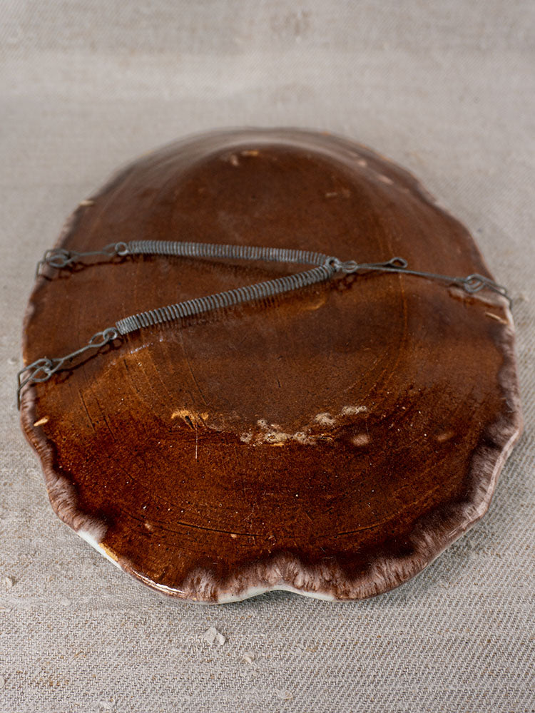Antique French faience platter upside down on neutral fabric, showing two metal hanging wires across its glossy brown ceramic back.