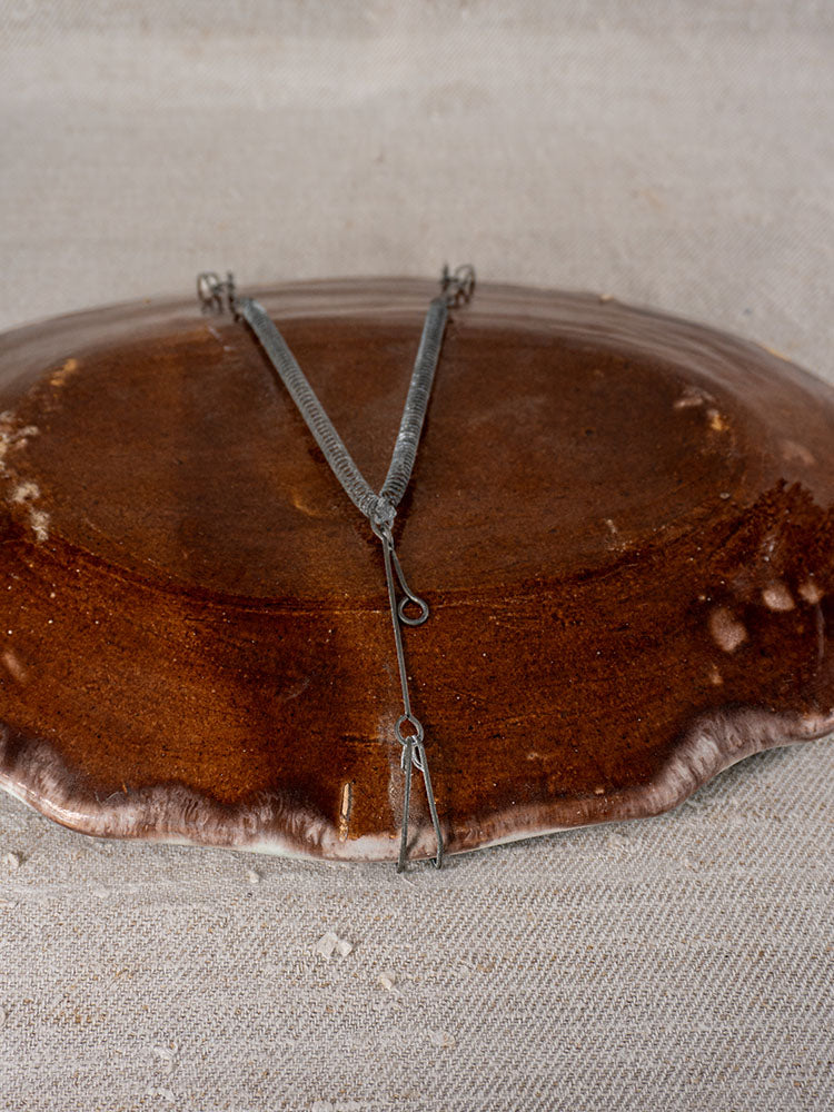 Back of antique French faience platter with metal wall hanger, displayed on textured beige fabric surface.