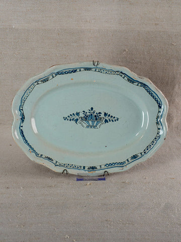 Oval French faience platter with scalloped edges, blue floral basket motif, set against beige textured fabric.