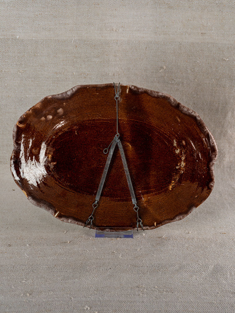 Antique French faience platter, Rouen Cul Noir, brown glazed underside with hanging wires, on textured beige fabric.