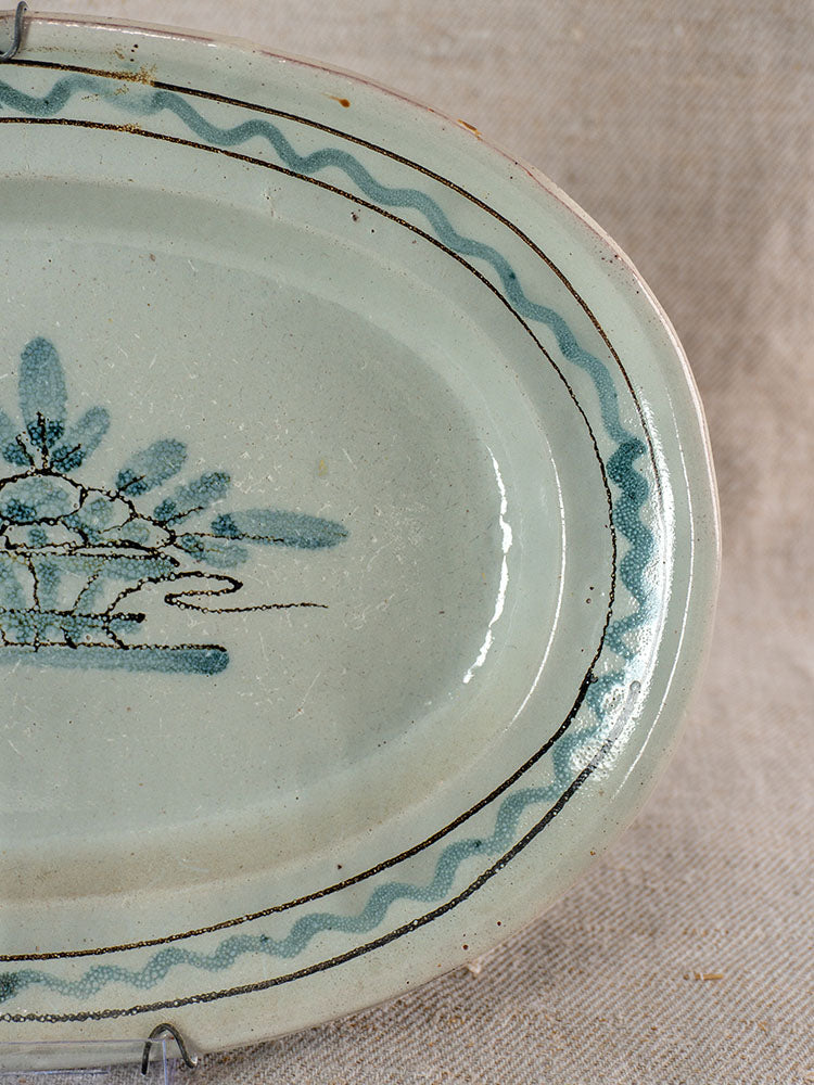 Antique French faience platter with blue-green glaze, hand-painted florals, wavy rim on beige fabric background.