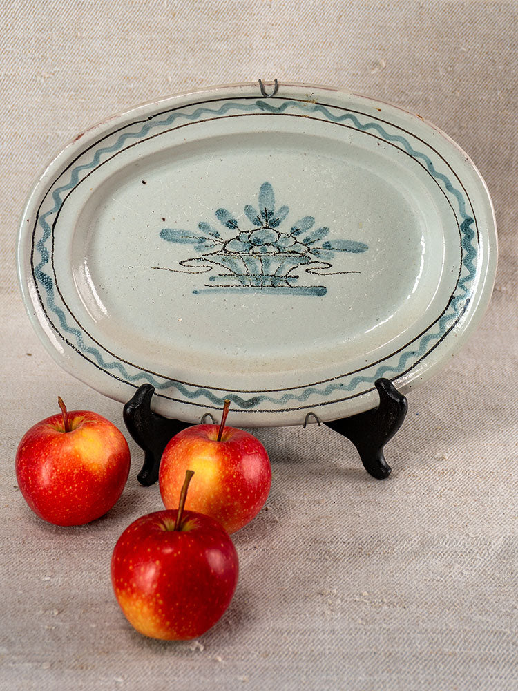 Antique French faience platter with blue floral design on black stand beside three red apples on beige fabric.