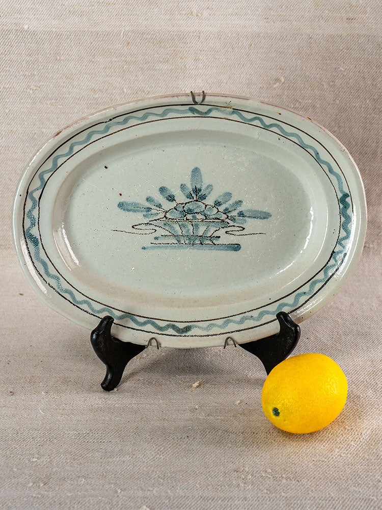 Antique French faience Cul Noir platter from Forges les Eaux on black stand beside a yellow lemon and neutral fabric backdrop.