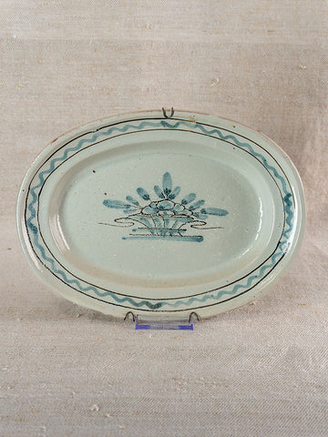 Antique French faience platter, Cul Noir, blue floral basket motif and wavy rim, on textured beige surface.