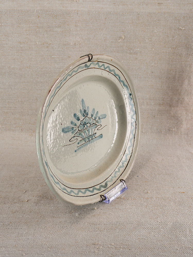 Antique French faience platter, round off-white ceramic with blue floral center, wavy rim, on beige textured fabric.