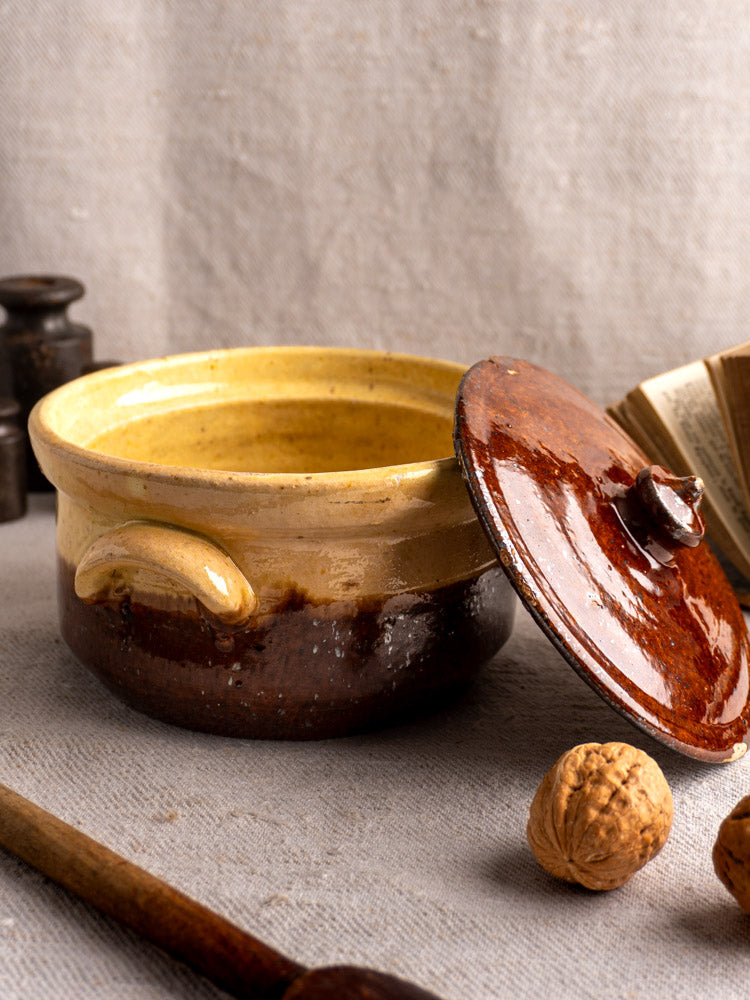 Antique French yellow ochre and brown glazed earthenware marmite with ajar lid on textured cloth, walnuts and utensils nearby.