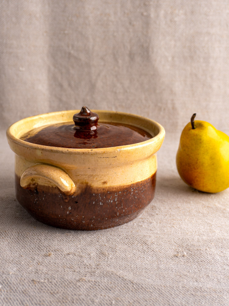 Antique French marmite, early 20th-century earthenware with yellow ochre and brown glaze, on beige surface with yellow pear.