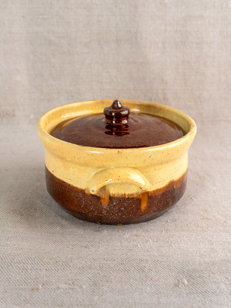 Antique French earthenware marmite with yellow ochre and brown glaze, displayed on textured beige fabric.