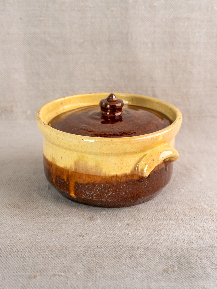 Antique French marmite in yellow ochre and brown glazed earthenware with lid on textured beige fabric.