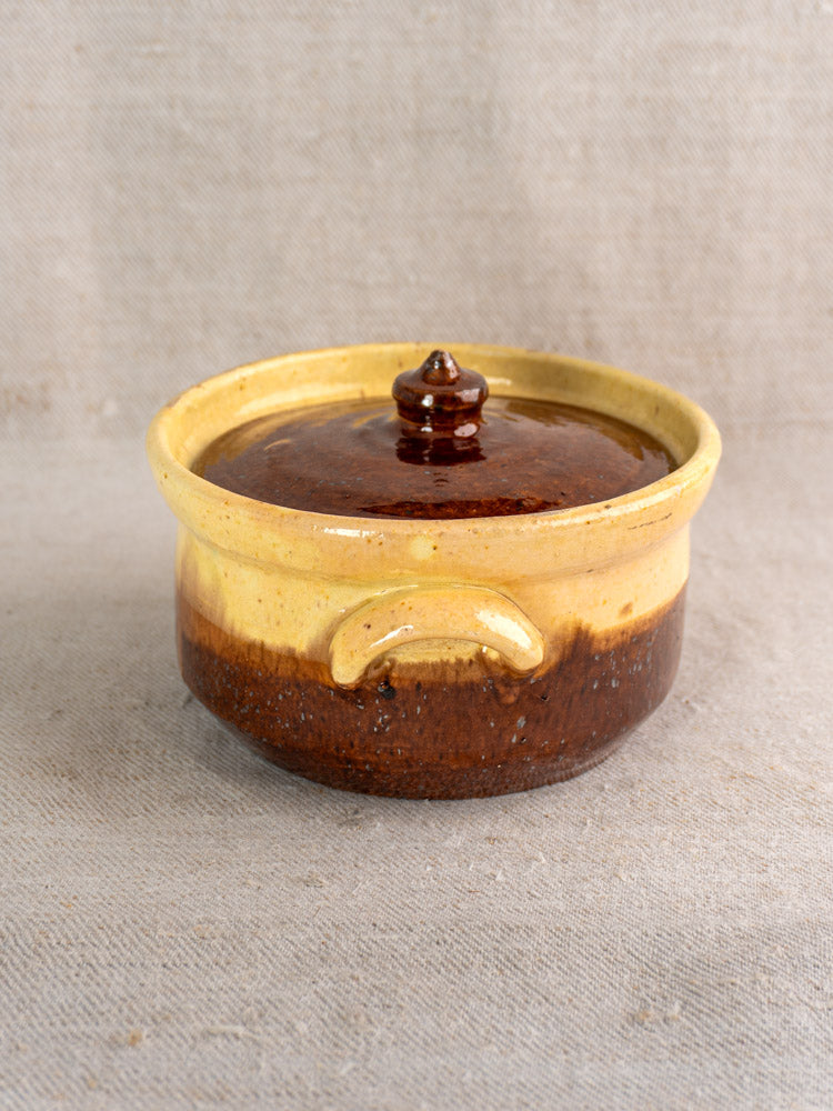 Early 20th-century French earthenware marmite with yellow ochre and brown glaze, handled lid, on beige fabric background.