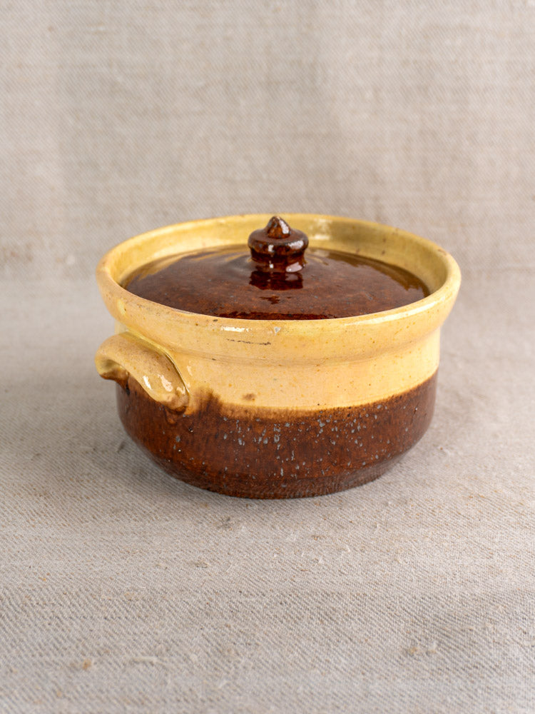 Antique French earthenware marmite with lid and side handle in ochre and brown glaze on beige fabric.